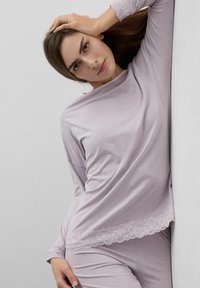 Frau lehnt an einer Wand, trägt ein hellviolettes langärmeliges Oberteil und eine Hose mit Spitzenbesatz, Hand auf dem Kopf ruhend.