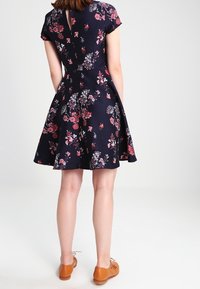 Šaty s květinovým potiskem v námořnické modři s přiléhavým korzetem a rozšířenou sukní, kratšími rukávy a detaily na zadním výstřihu ve tvaru klíčové dírky. Hnědé boty doplňují celý outfit.