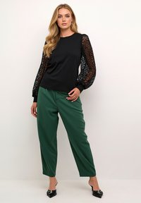 Black top with lace puff sleeves, paired with loose-fitting green trousers. Black pointed shoes complete the outfit. Smooth and textured fabrics.