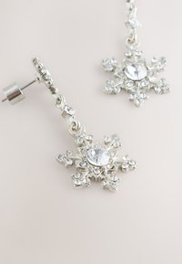 Srebrne naušnice u obliku pahuljica s prozirnim cirkonima na svijetloj pozadini, jedna leži ravno, a druga uspravno.