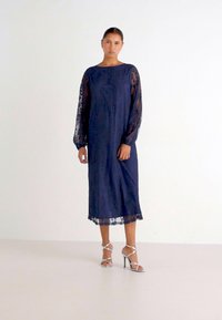 Vestido de renda azul-marinho com mangas longas transparentes, bainha até os tornozelos e padrões florais detalhados. Modelo a usar sandálias prateadas com tiras.
