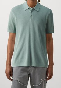 Hellgrünes Polo-Shirt mit kurzen Ärmeln aus strukturiertem Strick, mit Kragen und einer Knopfleiste mit drei Knöpfen. Kombiniert mit grauen Cargo-Shorts.
