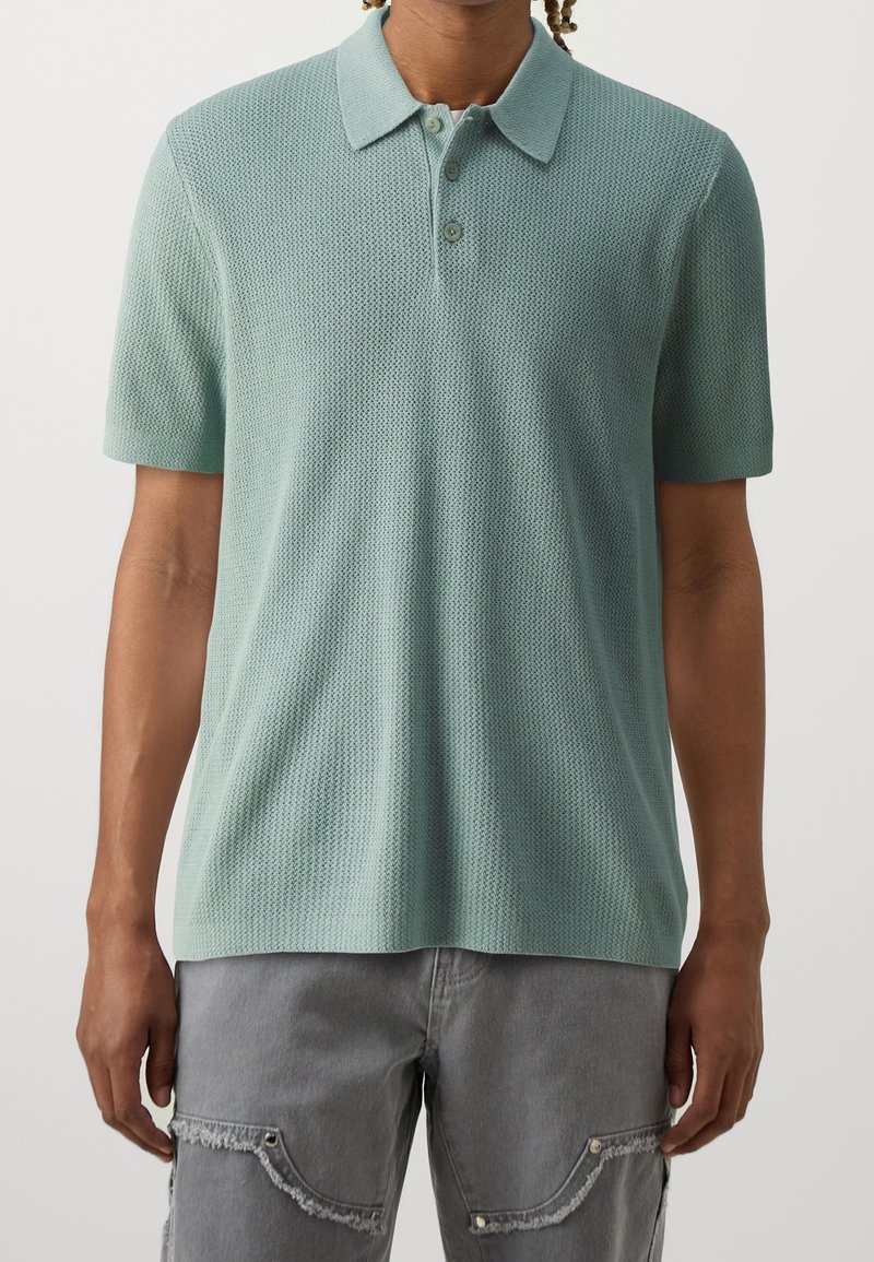 Hellgrünes Polo-Shirt mit kurzen Ärmeln aus strukturiertem Strick, mit Kragen und einer Knopfleiste mit drei Knöpfen. Kombiniert mit grauen Cargo-Shorts.