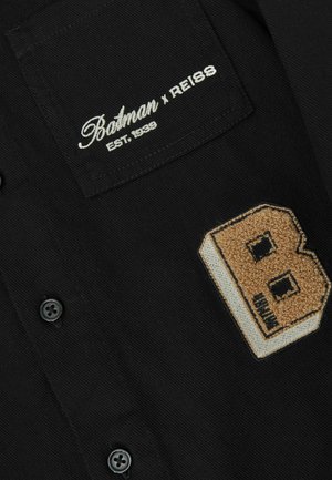 Juodas marškiniai su mygtukais, su tekstūruota rusva "B" pleistra ir baltu siuvinėtu logotipu "Batman x Reiss EST. 1939" ant kišenės.