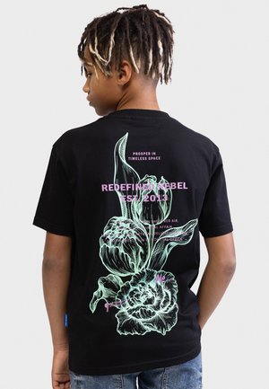 Jongen met dreadlocks met blonde punten draagt een zwart T-shirt met een grote pastelkleurige bloemenprint en roze tekst op de achterkant met de tekst "Redefined Rebel Est. 2013".