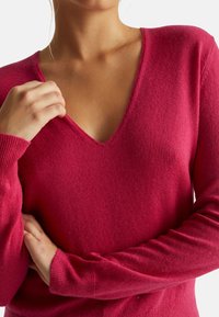 Donna che indossa un maglione rosso a scollo a V e maniche lunghe, che tiene il collo con una mano e ha le braccia incrociate sul petto.