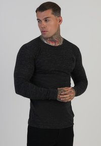 Fekete, hosszú ujjú, texturált anyagból készült póló, kerek nyakkivágással és testhezálló fazonnal. A modellen tetovált karok és kezek láthatók.