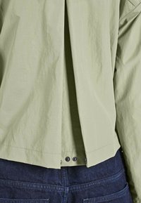 Ljusgrön, oversized skjorta med en texturerad yta, med en bakveck och tryckknappdetaljer vid fållen, bärs över mörk denim.