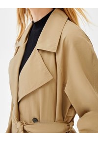Beżowy płaszcz trench z szerokim kołnierzem, z paskiem w talii i czarnymi guzikami. Nosić go na prążkowanej czarnej bluzce. Gładka faktura, casualowy design.