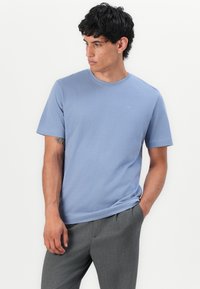 Himmelblaues Baumwoll-T-Shirt mit Rundhalsausschnitt und kurzen Ärmeln. Verfügt über ein kleines Emblem auf der Brust und wird mit grauen Hosen kombiniert.