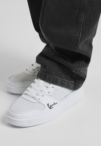 Valged perforatsiooniga tossud sileda pinnaga, millel on lamedad paelad ja külje peal äratuntav detail. Kombineeritud mustade teksapükstega.