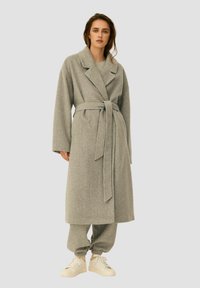 Szary długi płaszcz z klapami i paskiem, wykonany z miękkiego materiału. Stylizowany z pasującymi szarymi spodniami i białymi trampkami.