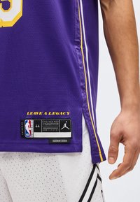 Κλείσιμο εικόνας σε μωβ φανέλα μπάσκετ NBA με το κείμενο «LEAVE A LEGACY» και ετικέτα μεγέθους 44, φορεμένη με λευκό κοντό παντελόνι με τρύπες και ορατό χέρι.