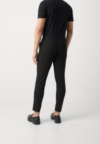 Černé kalhoty na míru se slim fit, vyrobené z hladkého materiálu. Mají pás s poutky na opasek a zadní kapsy. Nosené s černými botami.