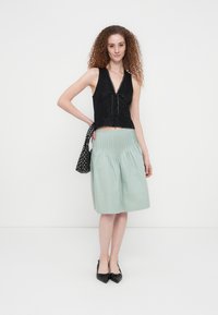 Μαύρο crop top από δαντέλα με V-λαιμόκοψη και μπροστινό δέσιμο, συνδυασμένο με φούστα σε μέντα πράσινο πλισέ. Το μοντέλο φοράει μαύρα παπούτσια με τακούνια και διακοσμητικές κορδέλες.