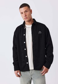 Schwarzes Hemd mit Knöpfen und einem strukturierten Fischgrätmuster, weißen Knöpfen und einem kleinen Logo auf der linken Brust. Über einem hellen Hemd getragen.