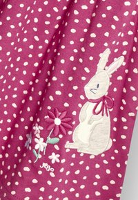 Tissu violet avec des pois blancs, mettant en avant un lapin blanc brodé et des fleurs colorées. Le texte "jojo" est visible près du motif.
