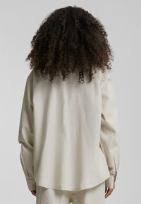 Beige langermet skjorte med avslappet passform, med en rett kant, knappede mansjetter og en glatt, myk tekstur. Vist fra baksiden.