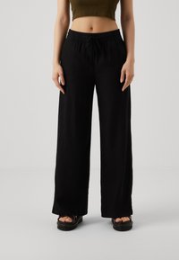 Pantalones anchos negros hechos de tejido ligero, que cuentan con una cinturilla elástica y cordón. Combinados con sandalias de plataforma oscuras.
