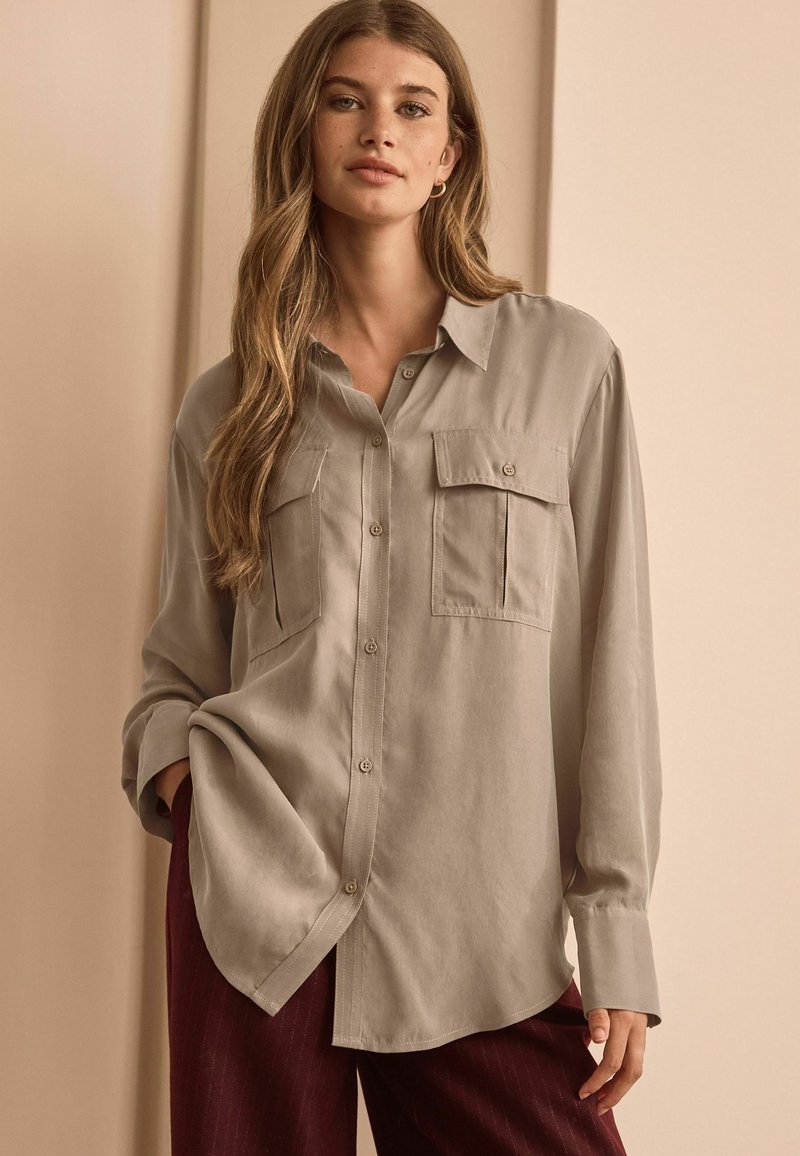 Light grey long-sleeve shirt with a button-down front, two chest pockets, and a relaxed fit. Smooth texture and curved hem.