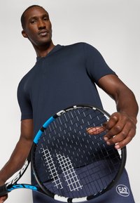 Polo azul-marinho de manga curta com um detalhe em botão, feito de um tecido texturizado. Uma pessoa segura uma raquete de ténis com detalhes em azul e preto.