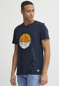 Marine bomulds t-shirt med et rundt grafisk design med en sol og bølger, rund halsudskæring, korte ærmer og et lille logo på nederste kant.