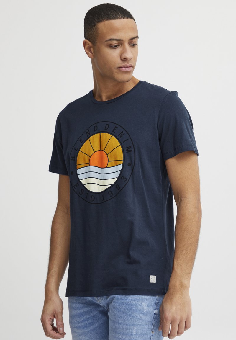 Marine bomulds t-shirt med et rundt grafisk design med en sol og bølger, rund halsudskæring, korte ærmer og et lille logo på nederste kant.