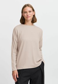 Light beige ribbed long-sleeve top with a round neckline, relaxed fit, and subtle texture, paired with black trousers.