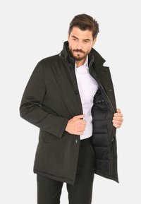 Cappotto verde scuro, resistente all'acqua, con colletto alto; presenta chiusura con bottoni e tasche laterali; rivestimento interno trapuntato visibile.