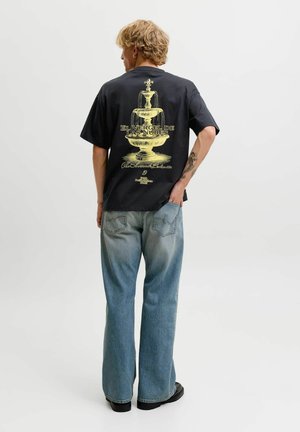 Personne aux cheveux blonds portant un t-shirt noir avec un graphisme de fontaine jaune et un texte, jean bleu clair, debout devant un fond uni.