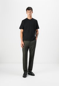 Camiseta de algodón negra combinada con pantalones verdes oliva. El modelo lleva zapatos de cuero negros, de pie contra un fondo blanco minimalista.