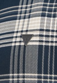 Kostkovaná látka v námořnické modré a béžové barvě. Na látce je výrazně umístěná trojúhelníková černá visačka s nápisem "GUESS?".