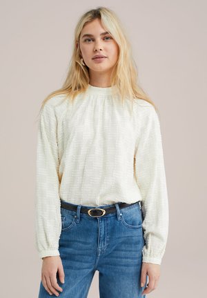 Blusa bianca con texture, maniche lunghe e collo alto, abbinata a jeans blu e una cintura nera con fibbia dorata.