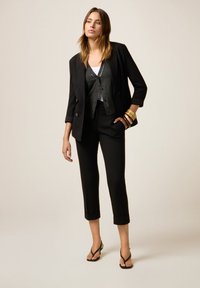 Femme portant un pantalon noir court, un blazer noir sur un cardigan gris foncé, un haut blanc, des sandales noires et des bracelets dorés, se tenant sur un fond beige.
