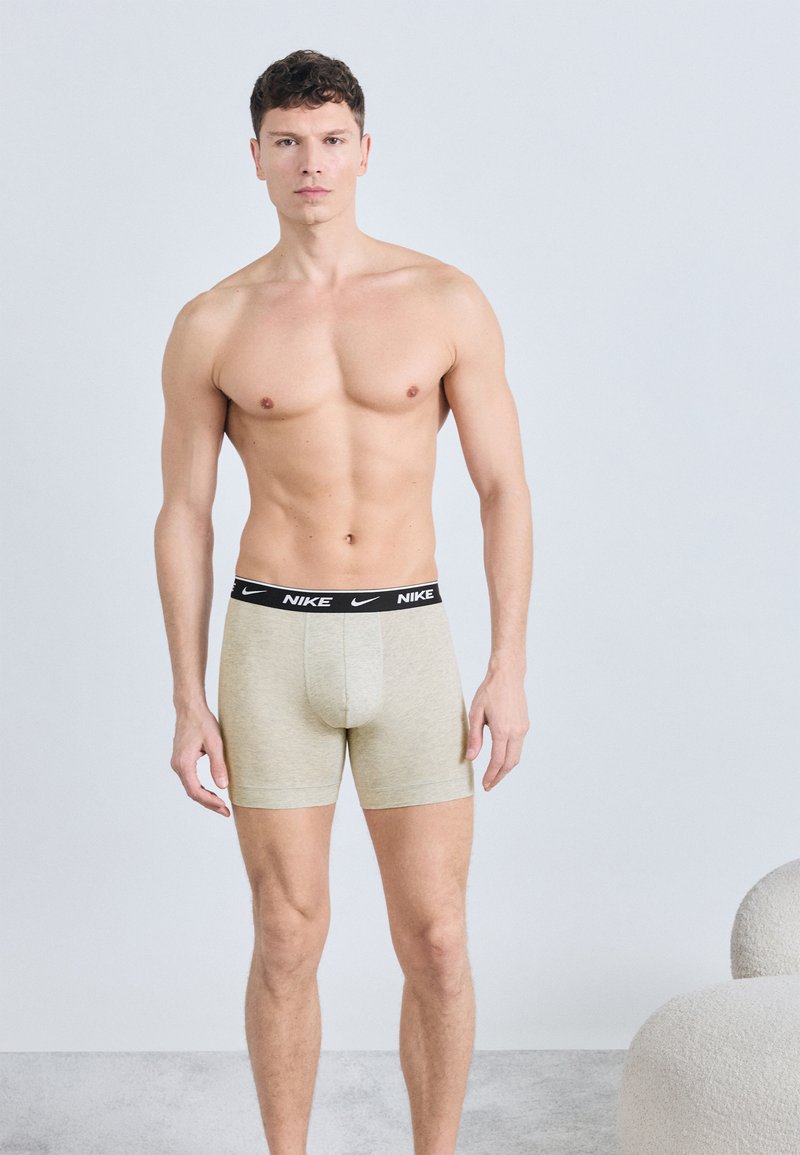 Μυώδης άνδρας που στέκεται μπροστά σε γκρι φόντο φορώντας μπεζ εσώρουχα της Nike με μαύρη ζώνη μέσης, χωρίς πουκάμισο, με ουδέτερη έκφραση.