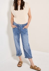 Kvinna som bär uppvikta ljusblå jeans, beige ärmlös topp och bruna sandaler med spänne, står med händerna i fickorna mot vit bakgrund.