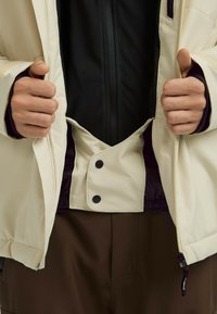 Lichtbeige jas met zwarte voering, vooraan met rits en kn closures. Voorzien van een elastische tailleband en een ritszak aan de onderkant. Bruine broek eronder.