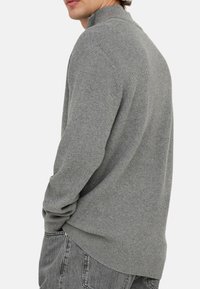 Grå ribbet sweater med høj krave, der har lange ærmer og en afslappet pasform. Stoffet er tekstureret og blødt, med slidser i siderne ved kanten.