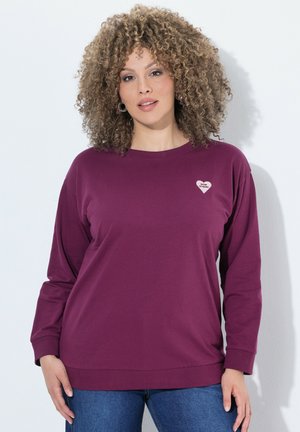 Kvinde med krøllet hår iført en lilla langærmet skjorte med en lille hjerteapplikation og blå jeans, stående mod en hvid baggrund.