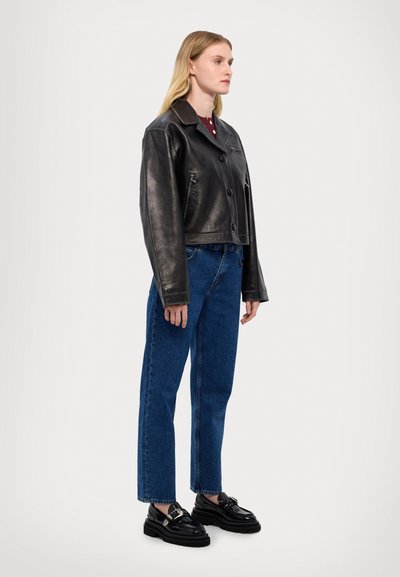 Schwarze Lederjacke mit Cropped-Schnitt, Knopfverschluss und breitem Kragen, kombiniert mit hochtaillierten blauen Jeans und schwarzen Plateauschuhen.