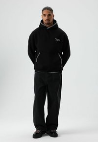 Zwarte capuchontrui met ritsen aan de voorkant en geborduurd ontwerp, gecombineerd met loszittende zwarte spijkerbroek en zwarte schoenen met een laag profiel.