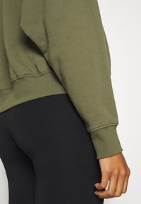 Gros plan d'une personne portant un haut à manches longues vert olive avec des poignets côtelés et un pantalon noir moulant.