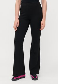 Pantalons évasés noirs en tissu extensible, dotés d'une taille haute. Associés à des chaussures de sport roses et noires pour le contraste.