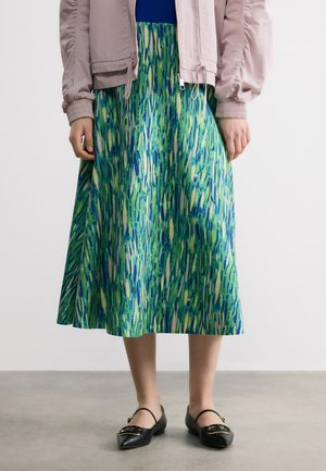 Personne portant une jupe midi à motifs verts et bleus, une veste rose clair, des ballerines noires pointues, debout sur un sol gris devant un mur blanc uni.