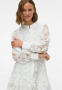 Weißes Spitzenkleid mit Blumenmustern, hohem Kragen und langen Ärmeln, mit gestuftem Saum und Knopfverschluss an der Vorderseite.