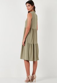Sleeveless olive-green dress with a tiered design, softly textured fabric, and a high neckline, paired with nude high-heeled sandals.