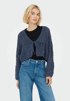 Blå hæklede cardigan med åbenmønstret design, cropped snit og bindelukning foran, båret over en sort bluse og parret med blå jeans.