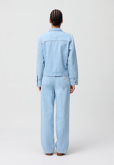 Veste en denim bleu clair associée à un jean ample assorti. La veste possède un col et des poignets à boutons. Poches arrière sur le jean avec des détails de couture discrets.