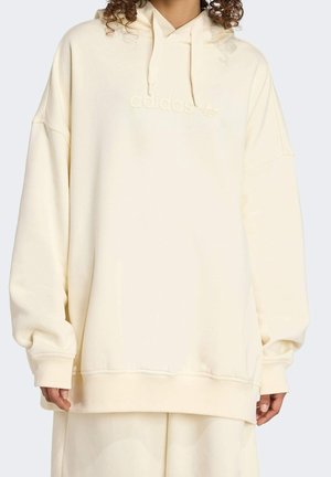 Personne portant un sweat à capuche crème oversize Adidas avec logo embossé et pantalon ample assorti sur fond uni.