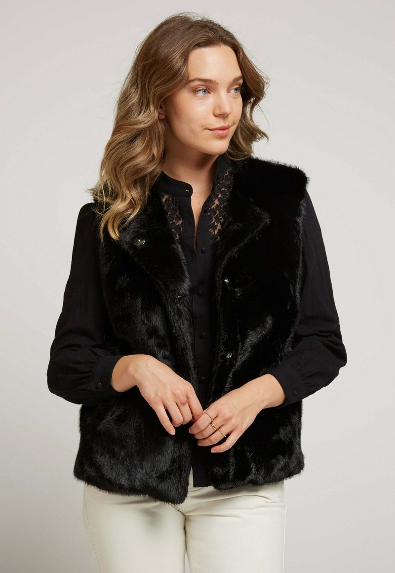 Gilet en fausse fourrure noire avec une texture douce, doté d'un col et de détails matériels subtils, associé à une blouse noire à boutons et à un pantalon crème.